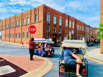 Tourists ride golf carts past brick buildings at a stop sign on a sunny downtown street.
