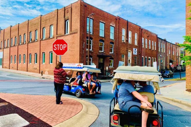 Tourists ride golf carts past brick buildings at a stop sign on a sunny downtown street.
